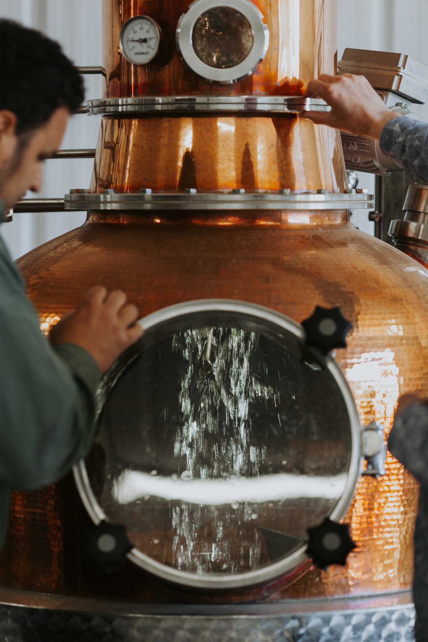 Two people working with a vintage copper pot still in a distillery setting, made by Mueller